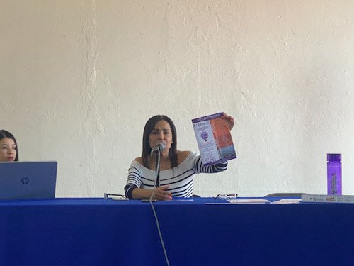 Mujer con una hoja en la mano sentada y al frente una mesa color morado