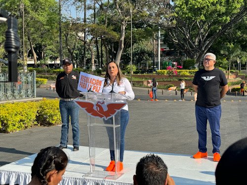 Mujer con tenis naranjas y blusa blanca, acompañada de dos hombres más con los logos de Movimiento Ciudadano