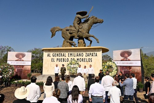 Estatua de Emiliano Zapata ofrenda floral y varias personas frente ala ofrenda